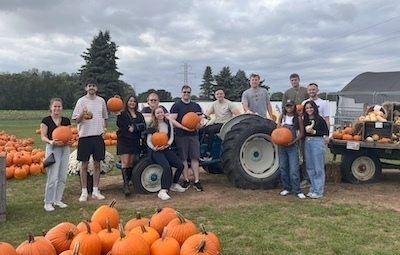 A group of students poses with pumpkins at a pumpkin patch. Some students hold pumpkins while others stand near a blue tractor. Large piles of pumpkins surround the group, creating a festive fall scene.