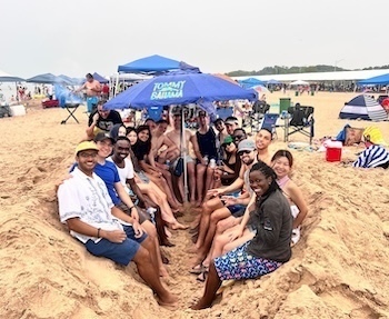 A group of students smiles for a photo while sitting together in a shallow sandy pit on a crowded beach under a Tommy Bahama umbrella.
