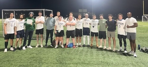 The ESTEEM soccer team poses for a photo on a turf field at night. The players wear white t-shirts with "ESTEEM" and individual numbers printed on them. A soccer goal is visible in the background.