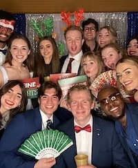 A group of smiling students pose for a photo in front of a blue and white backdrop. Some wear festive holiday attire like Santa hats and reindeer antlers.