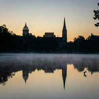 Golden Dome and Basilica of the Sacred Heart silhouette against a gradient orange and blue dawn sky. Their reflections appear in misty St. Mary's Lake, where a duck swims.