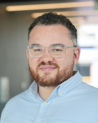 Light-skinned man with dark curly hair, reddish beard, clear glasses, and light blue shirt smiles gently.